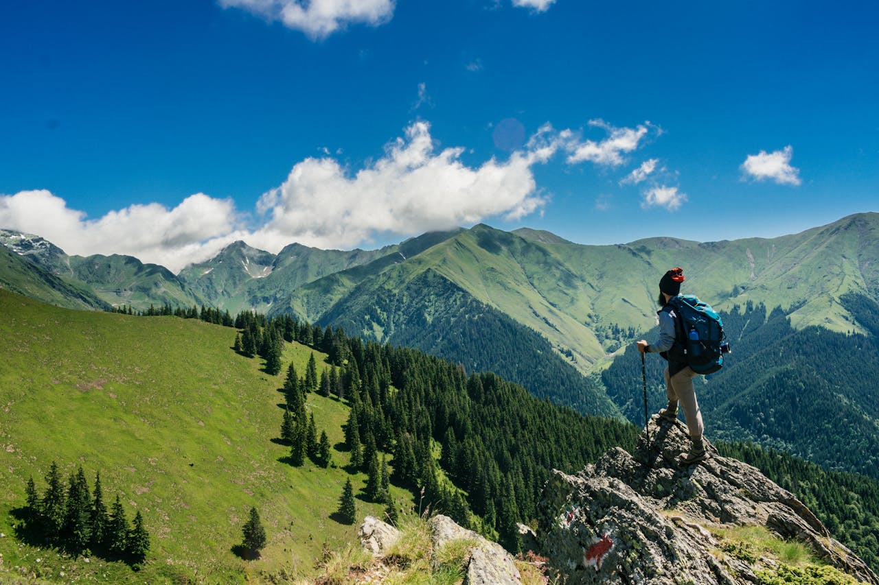 who-we-are Adventurous hiker enjoying breathtaking views of lush green mountains in Romania under a clear blue sky.