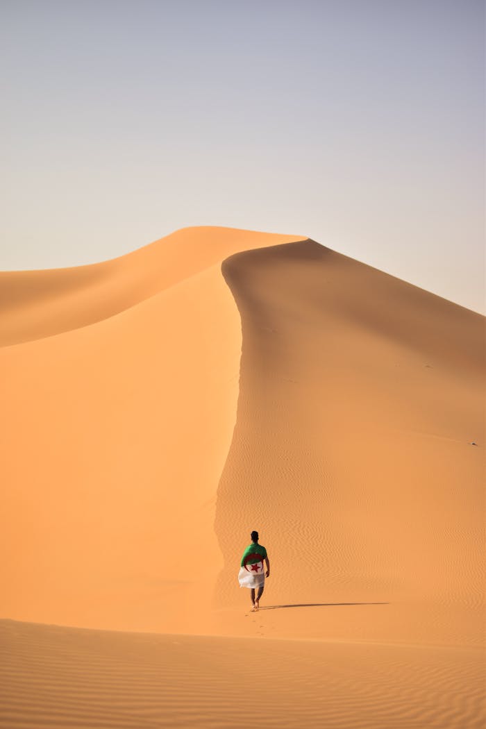 our-story A lone traveler walking over the vast, golden sand dunes of the Sahara Desert in Algeria.